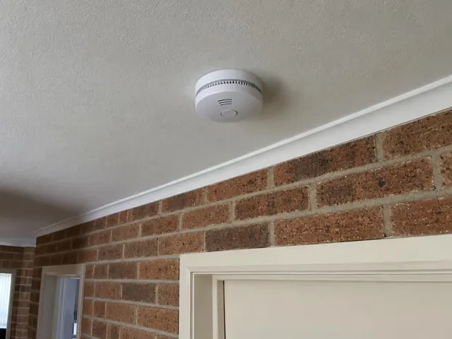 Ceiling-mounted smoke alarm above a brick hallway wall and doorway in Edgeworth